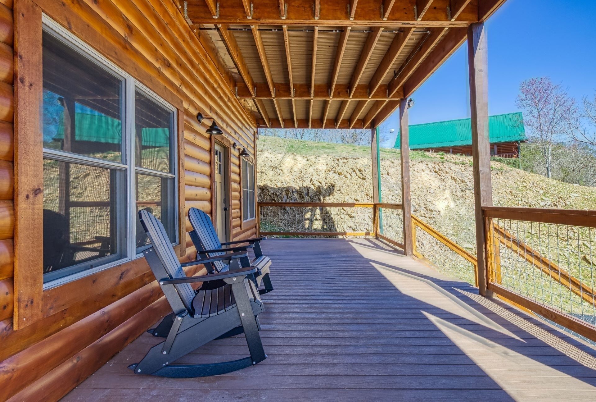 A wooden cabin with a covered porch, two rocking chairs, and a view of a green building in the distance.
