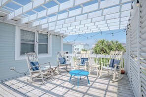 Una terraza cubierta al aire libre con sillas de madera y una mesita, adosada a una casa de fachada blanca.