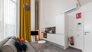 A modern living room with a sofa, a flat-screen TV, and a small table. A kitchen is visible with white cabinets and a fire extinguisher.
