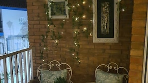 A balcony with two white metal chairs, a small table, and a potted plant. There are string lights and framed pictures on the brick wall.