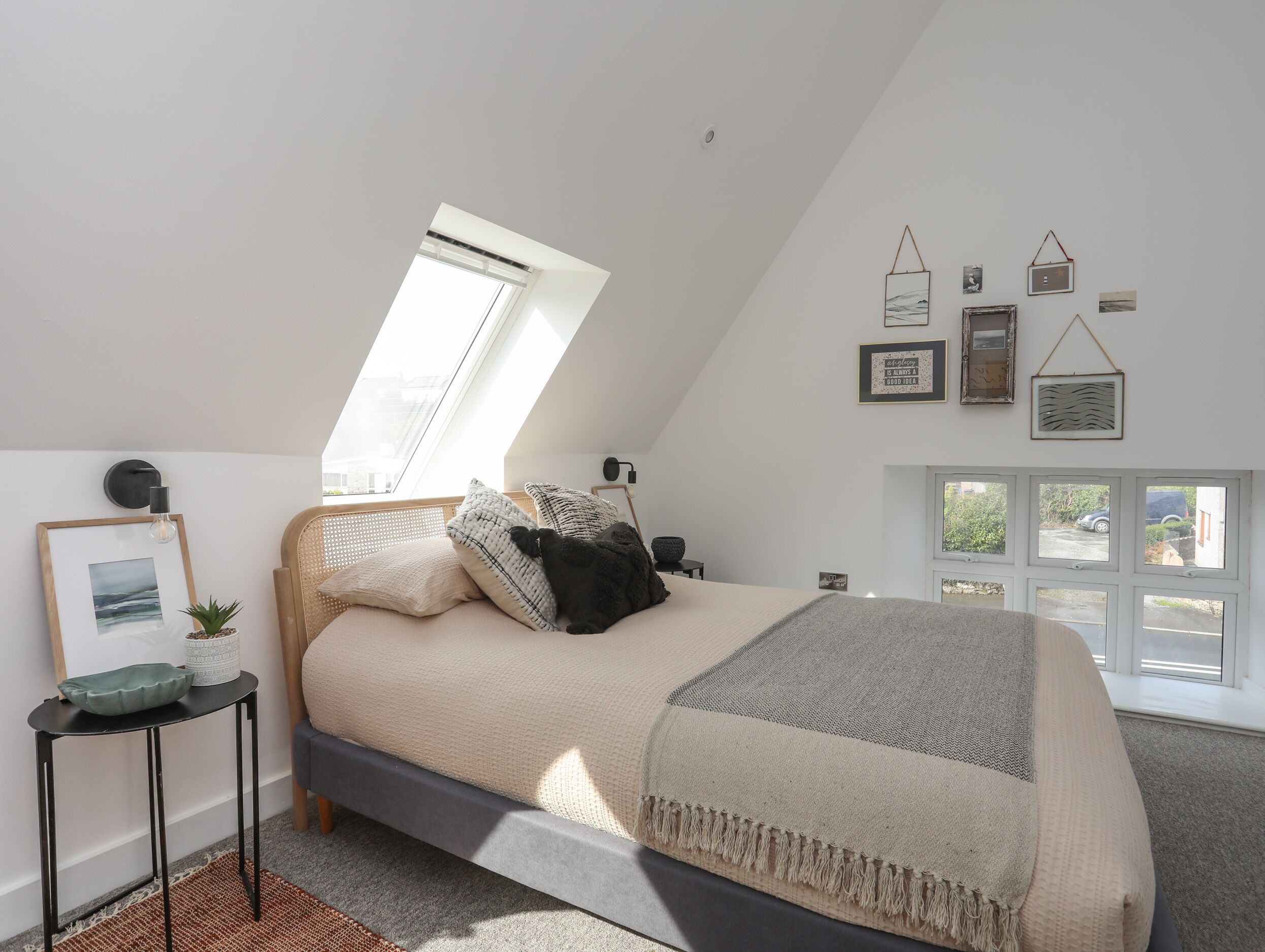 Ein Schlafzimmer mit schräg verlaufender Decke, einem Bett mit grauem Kopfteil, einem kleinen Tisch mit Pflanze und einem Fenster mit Jalousien.