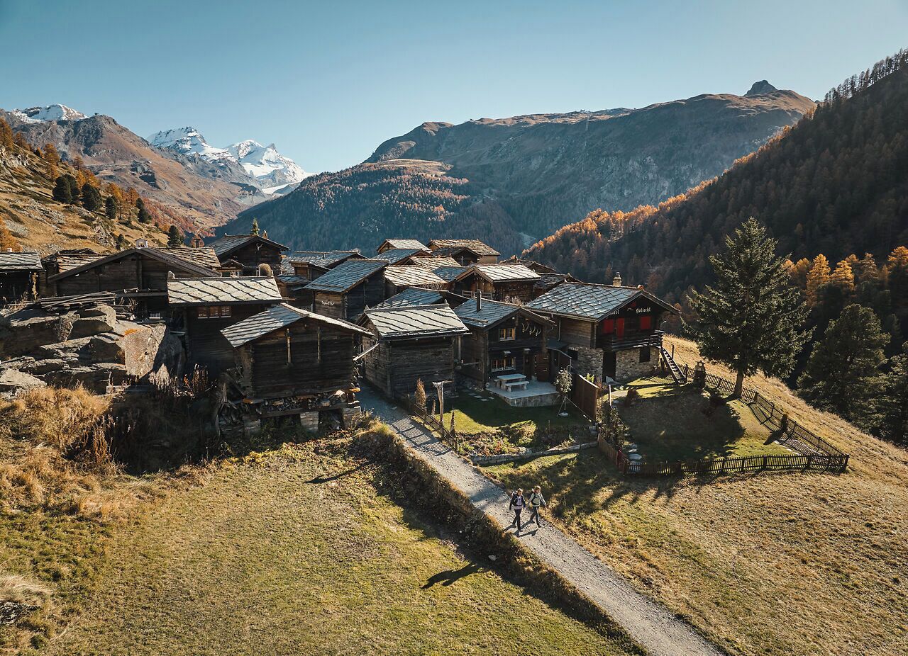 A mountain village with rustic wooden buildings, a clear sky, and snow-capped peaks in the background.