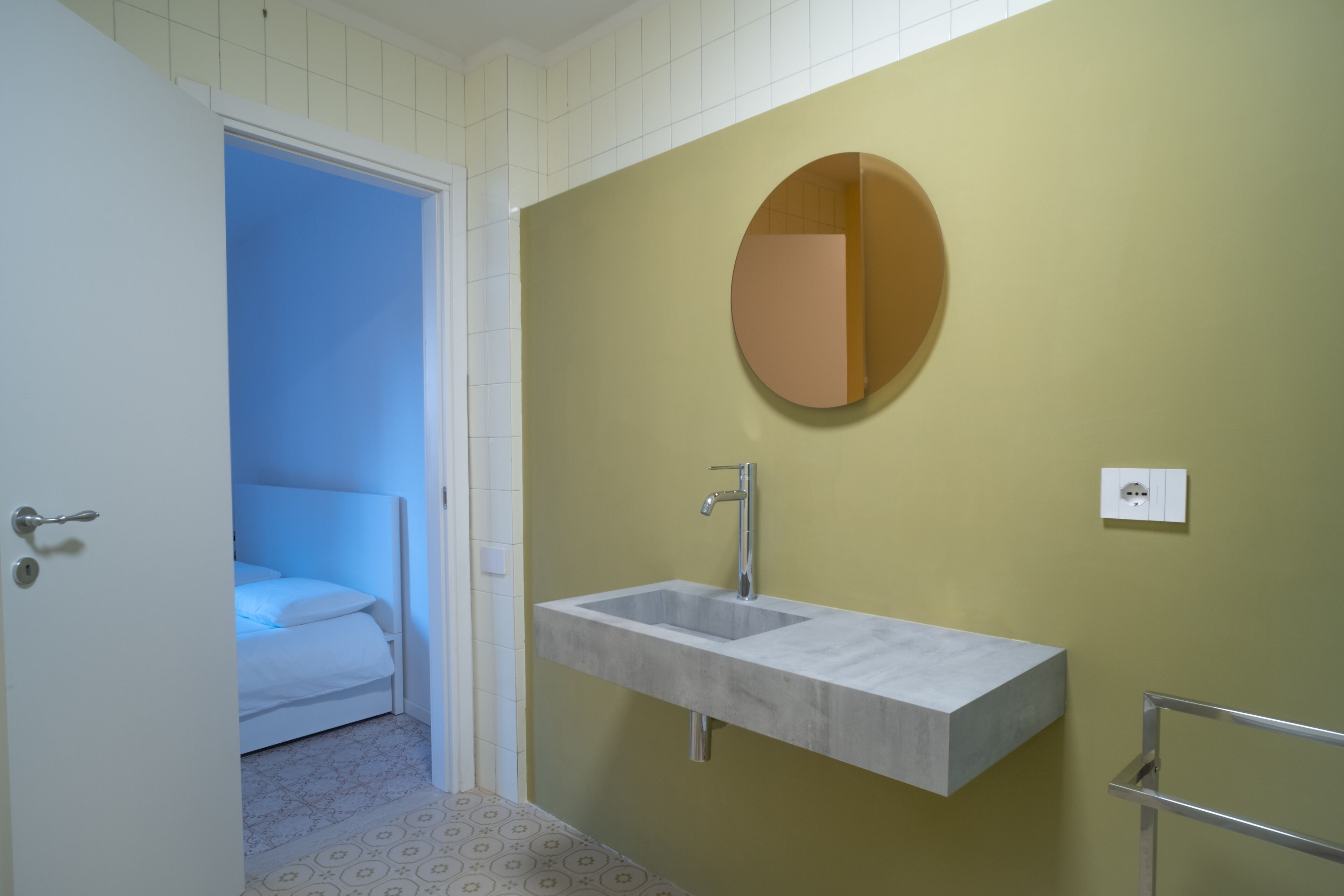 Ein Badezimmer mit einem weißen Waschbecken, einem runden Spiegel und einer Tür, die zu einem Schlafzimmer führt.