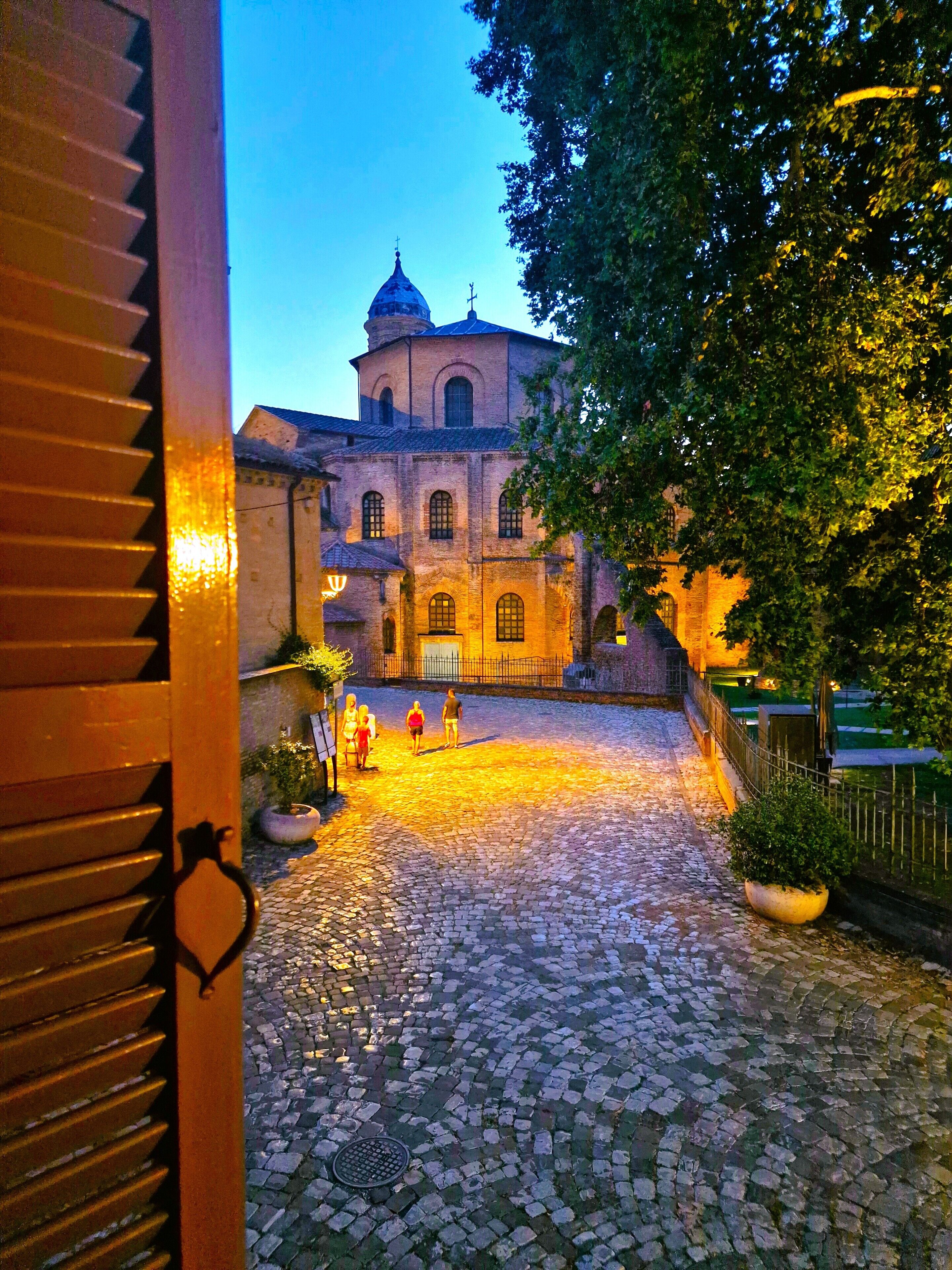 A cobblestone courtyard with a building featuring arched windows and a dome, surrounded by trees and a fence.