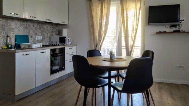 Una cocina moderna con armarios blancos, un microondas y una estufa. Hay una zona de comedor con una mesa de madera y cuatro sillas. Un televisor está montado en la pared sobre la mesa.