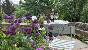 En have med hvide metal stole og et bord med glasplade, omgivet af lilla blomster og grønt.
