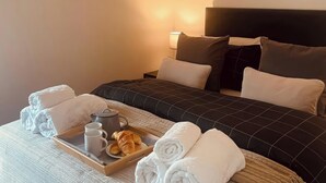 En pænt redt seng med et mørkt tæppe, et sengebord med en lampe, og et bakke med håndklæder, croissanter og et tekopper.