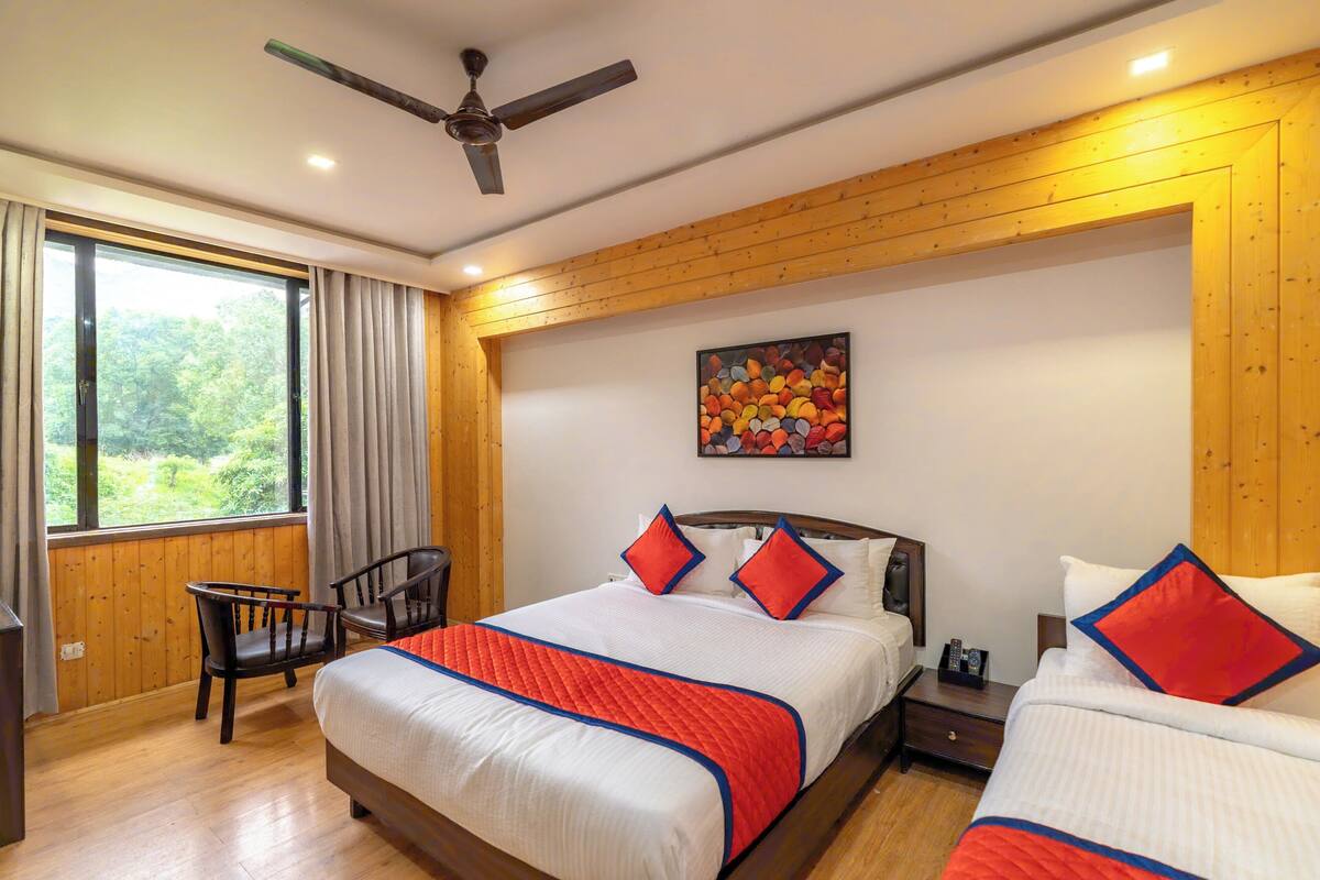 A hotel room with two beds, a ceiling fan, a large window with curtains, a painting on the wall, and a wooden floor.
