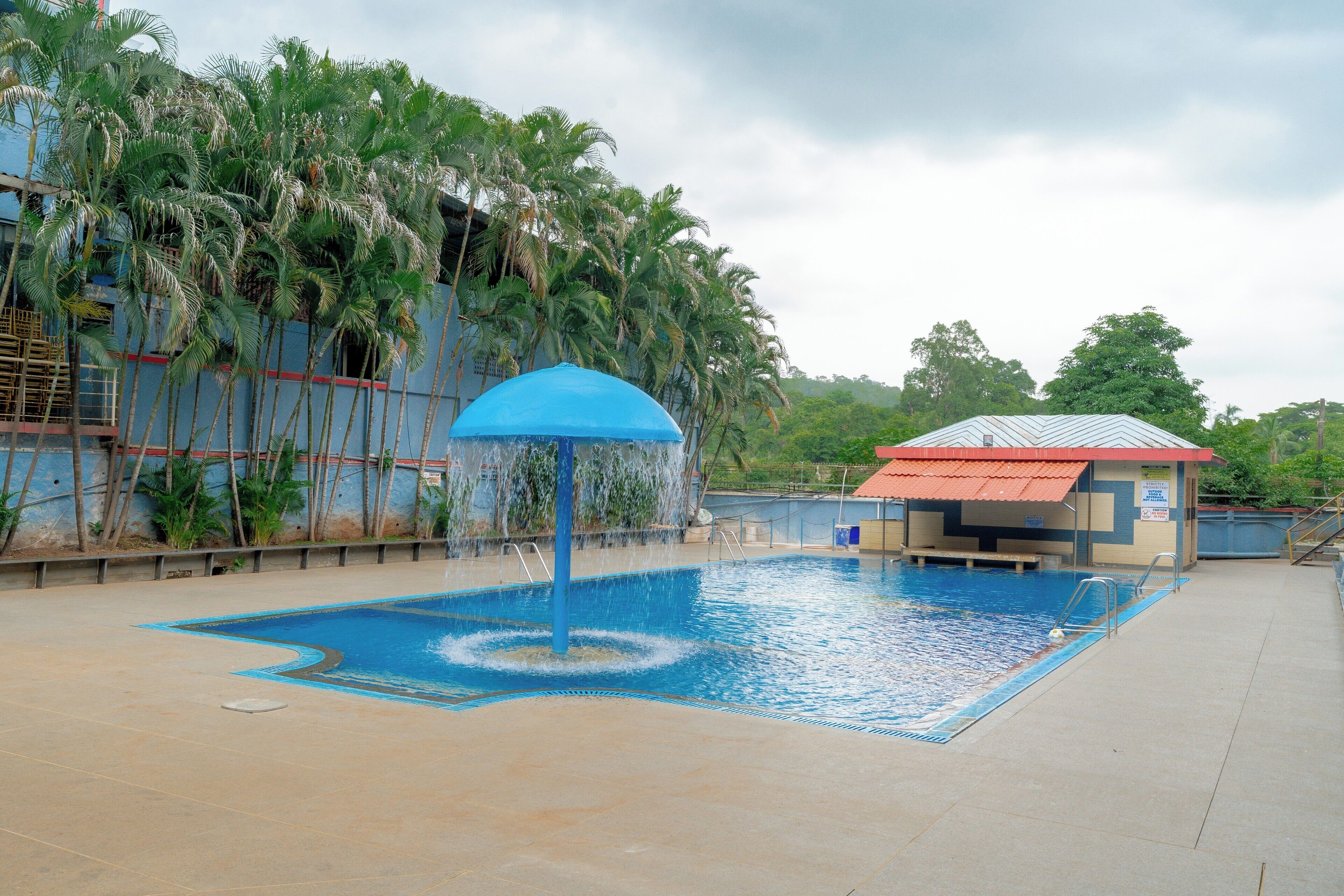 Ein Swimmingpool mit einer blauen Wasserspiels, umgeben von Palmen und einem Gebäude mit rotem Dach.