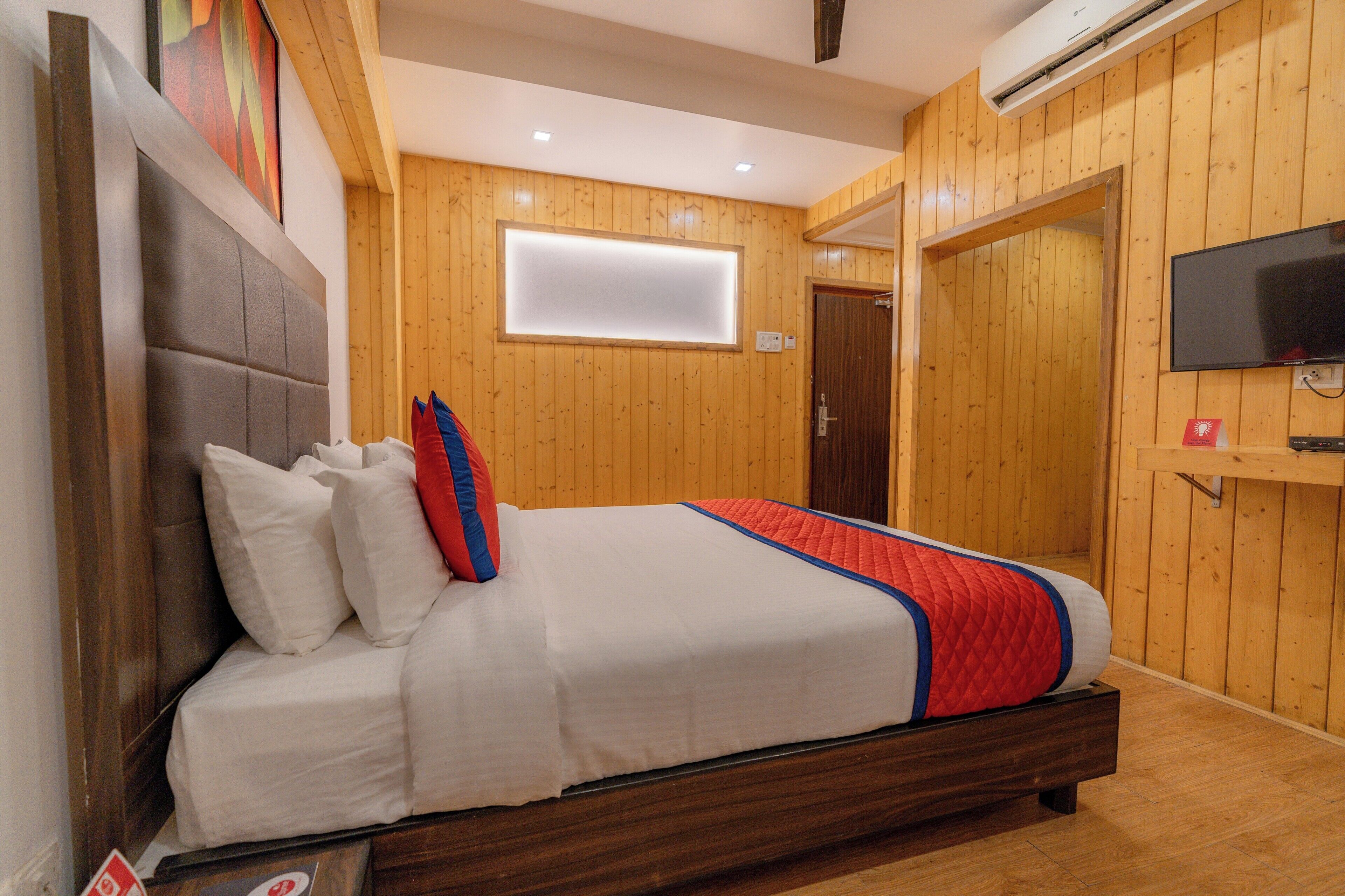 A bedroom with a wooden headboard, a bed with white bedding and a red and blue quilt, a flat-screen TV mounted on the wall, and a wooden door.