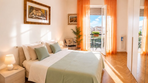 A bedroom with a bed, nightstand, lamp, two framed pictures, a potted plant, and a view of the outside.