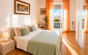Una camera da letto con un letto, un comodino, una lampada, due quadri appesi, una pianta in vaso e vista sull'esterno.