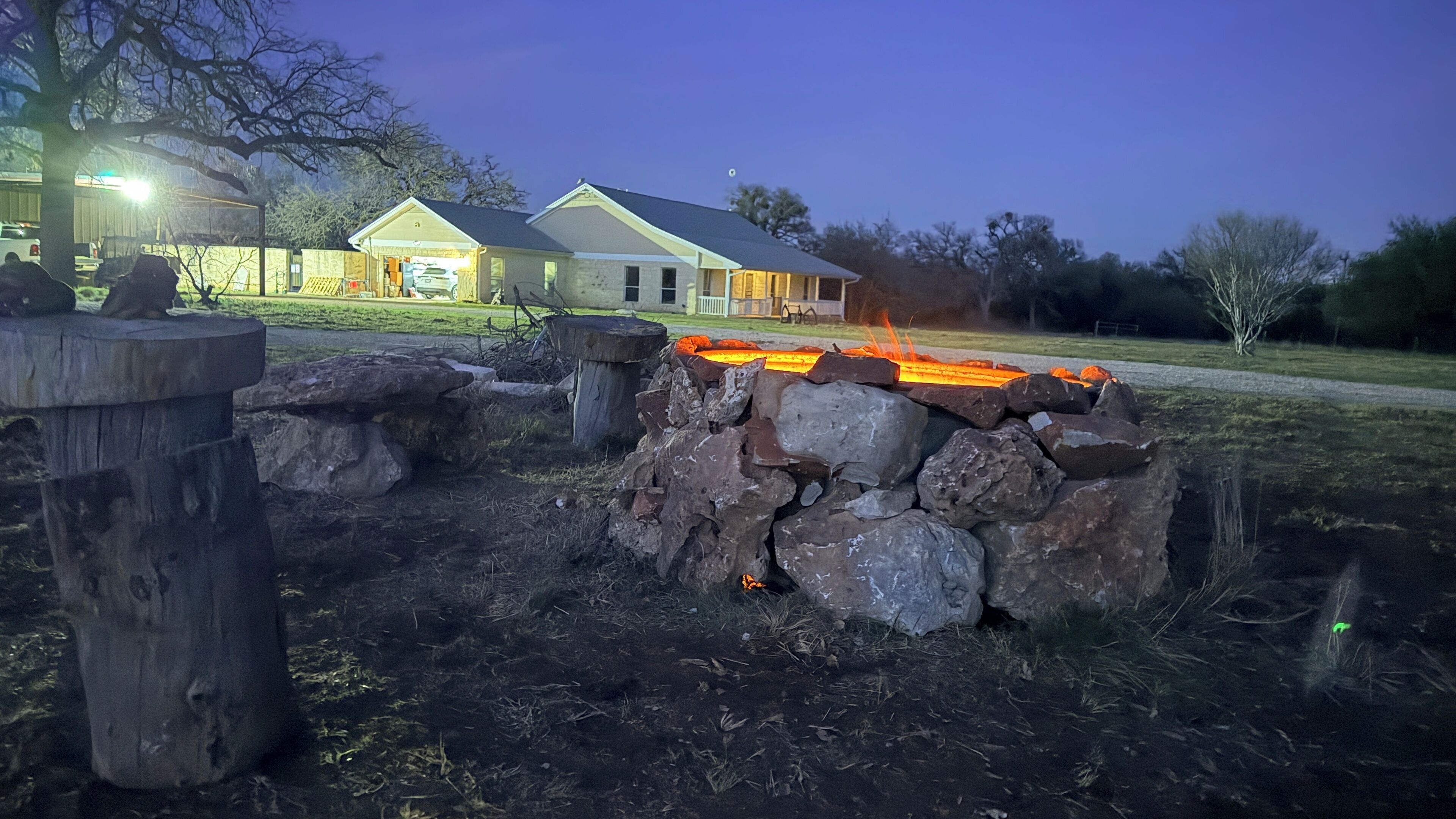 A stone fire pit with a fire burning, surrounded by rocks and a wooden post. In the background, there is a house with a garage and a lawn.