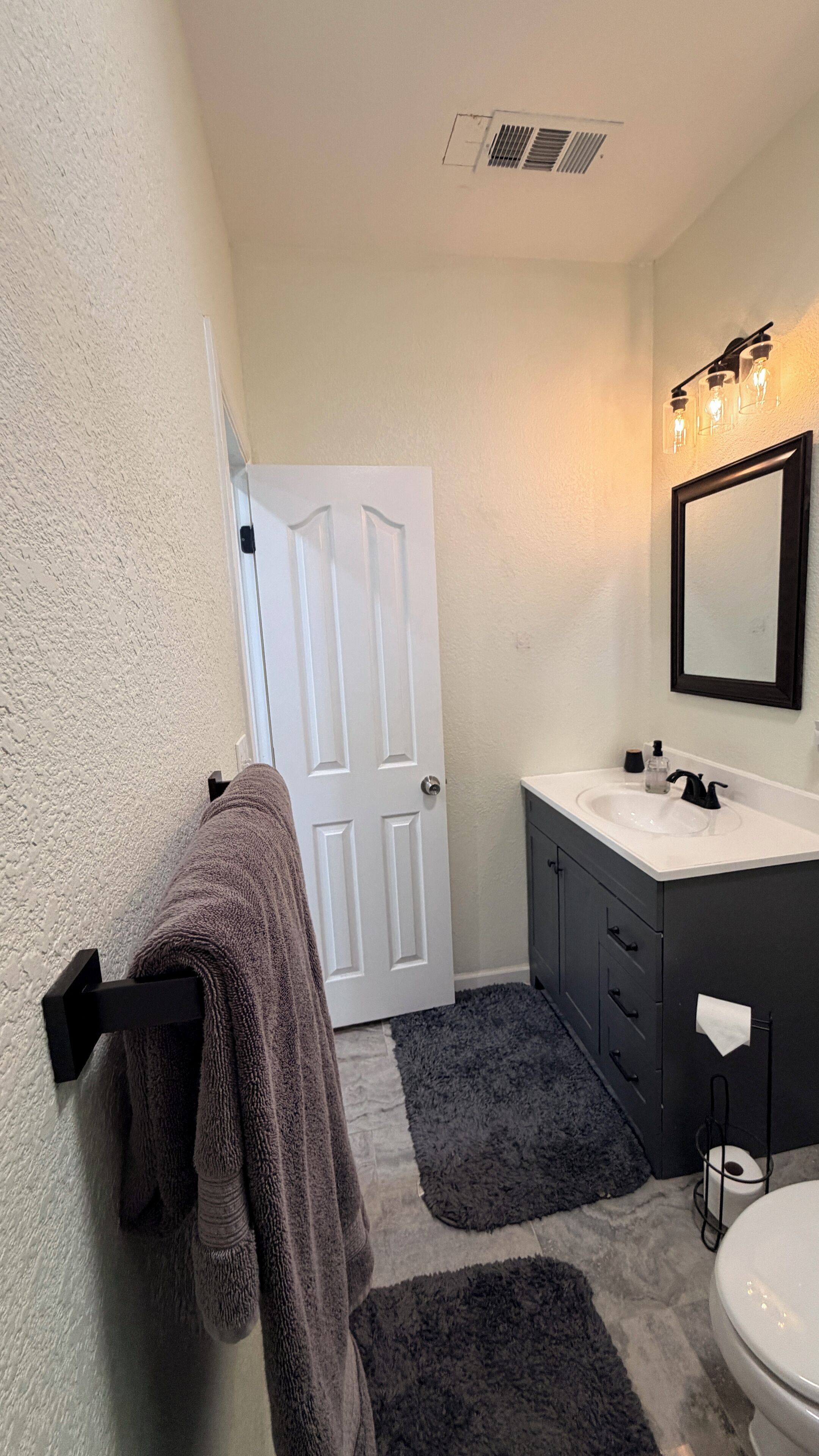 Un baño con una puerta blanca, un lavabo con espejo y un tapete negro.