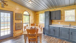 A wooden cabin interior with a dining table, chairs, a kitchen with grey cabinets, and a large window. - Melbourne Home w/ Fire Pit on the White River! (Melbourne)