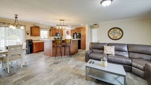 A spacious living area with a brown leather sofa, a dining table with chairs, and a kitchen area with wooden cabinets and a bar stool. - Efficiently Equipped Lewis Home: Deck & Gas Grill! (Lewis)