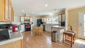 Una cocina amplia con armarios de madera, un comedor con sillas, un bote de basura de acero inoxidable y un ventanal que proporciona luz natural.