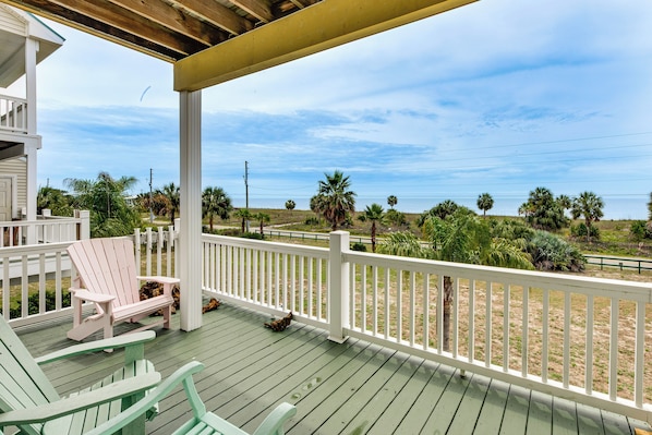 A wooden deck with white railings, two green Adirondack chairs, and a view of a landscaped area with palm trees.