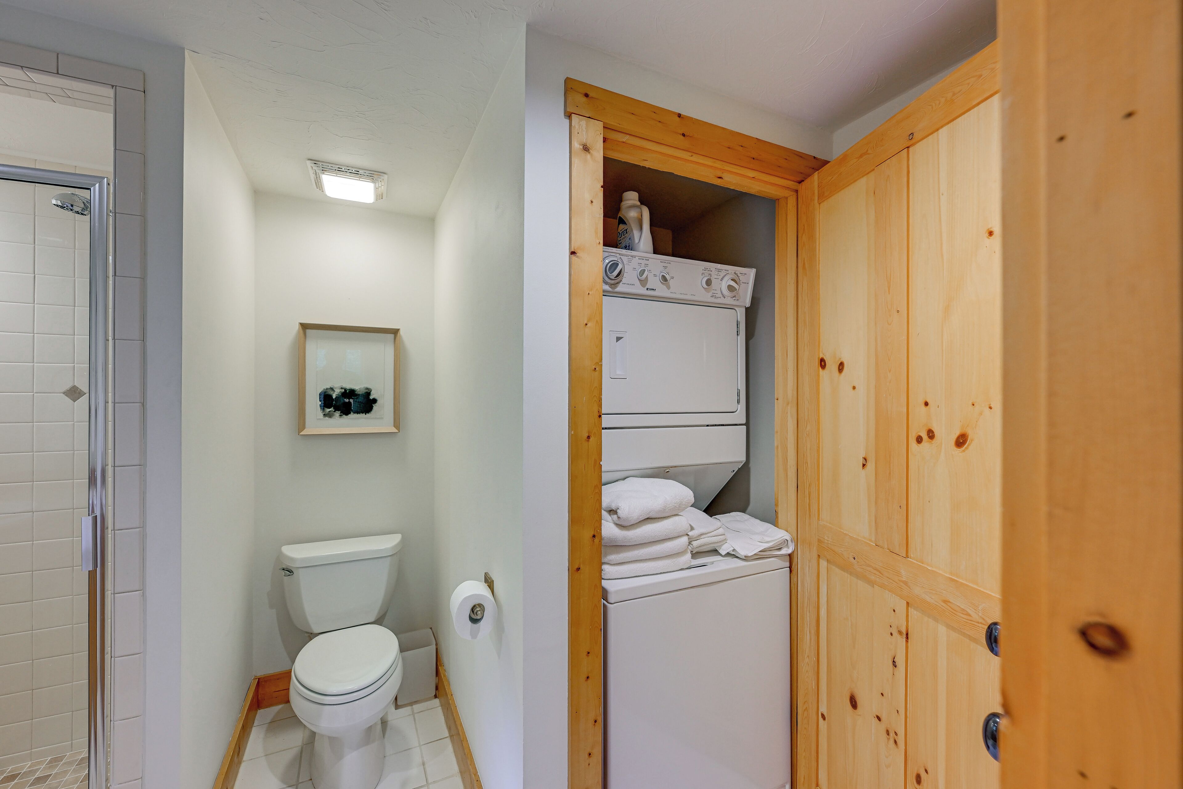 Un bagno con il water, un quadro appeso al muro e una porta in legno che conduce a un'area lavanderia con lavatrice e asciugamani impilati.