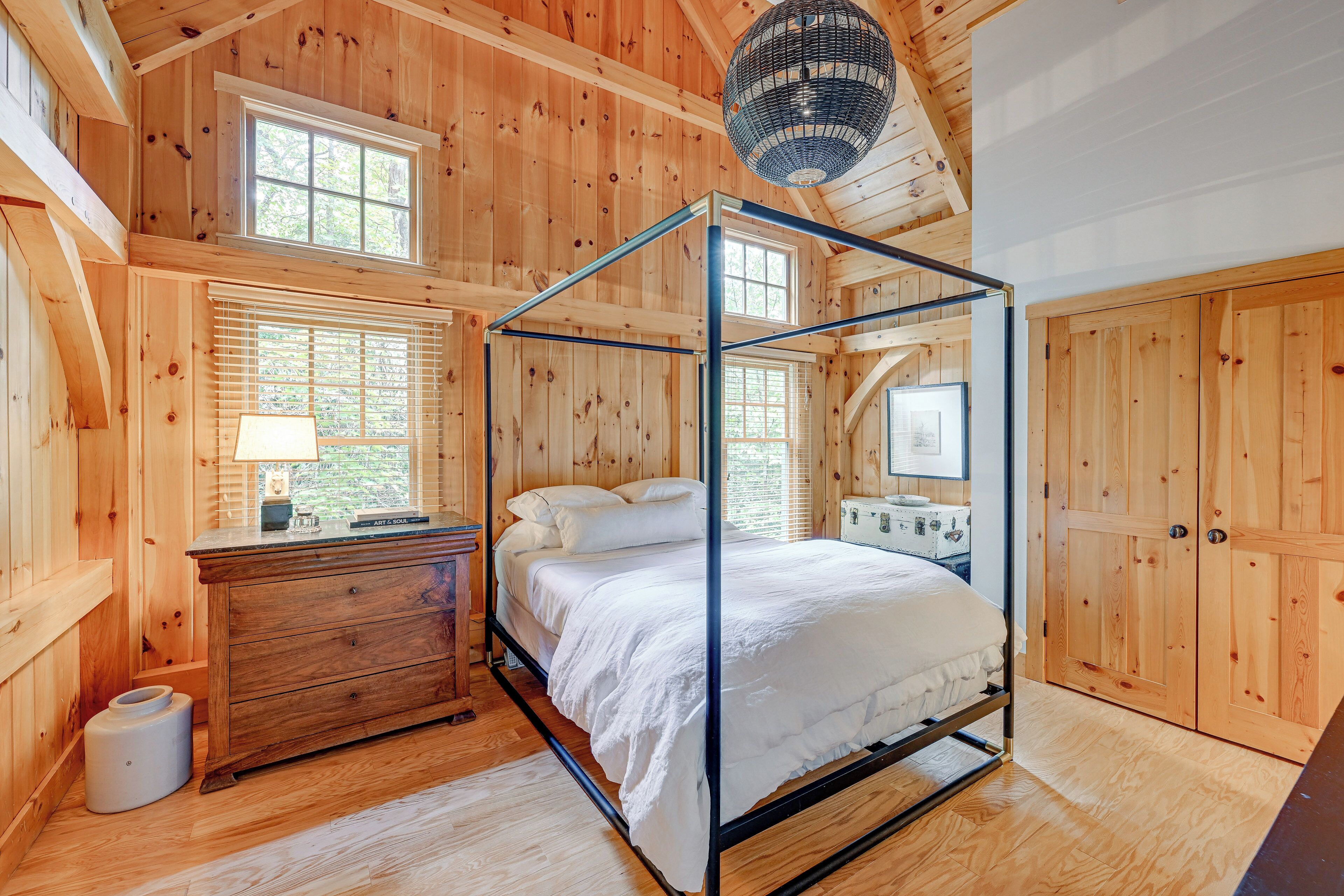 A wooden cabin bedroom with a four-poster bed, a dresser, and a window with blinds.