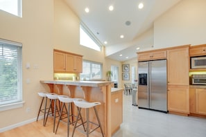 A modern kitchen with wooden cabinets, a stainless steel refrigerator, and a bar area with stools. - Cozy Waldport Vacation Rental: Steps From the Bay! (Waldport)