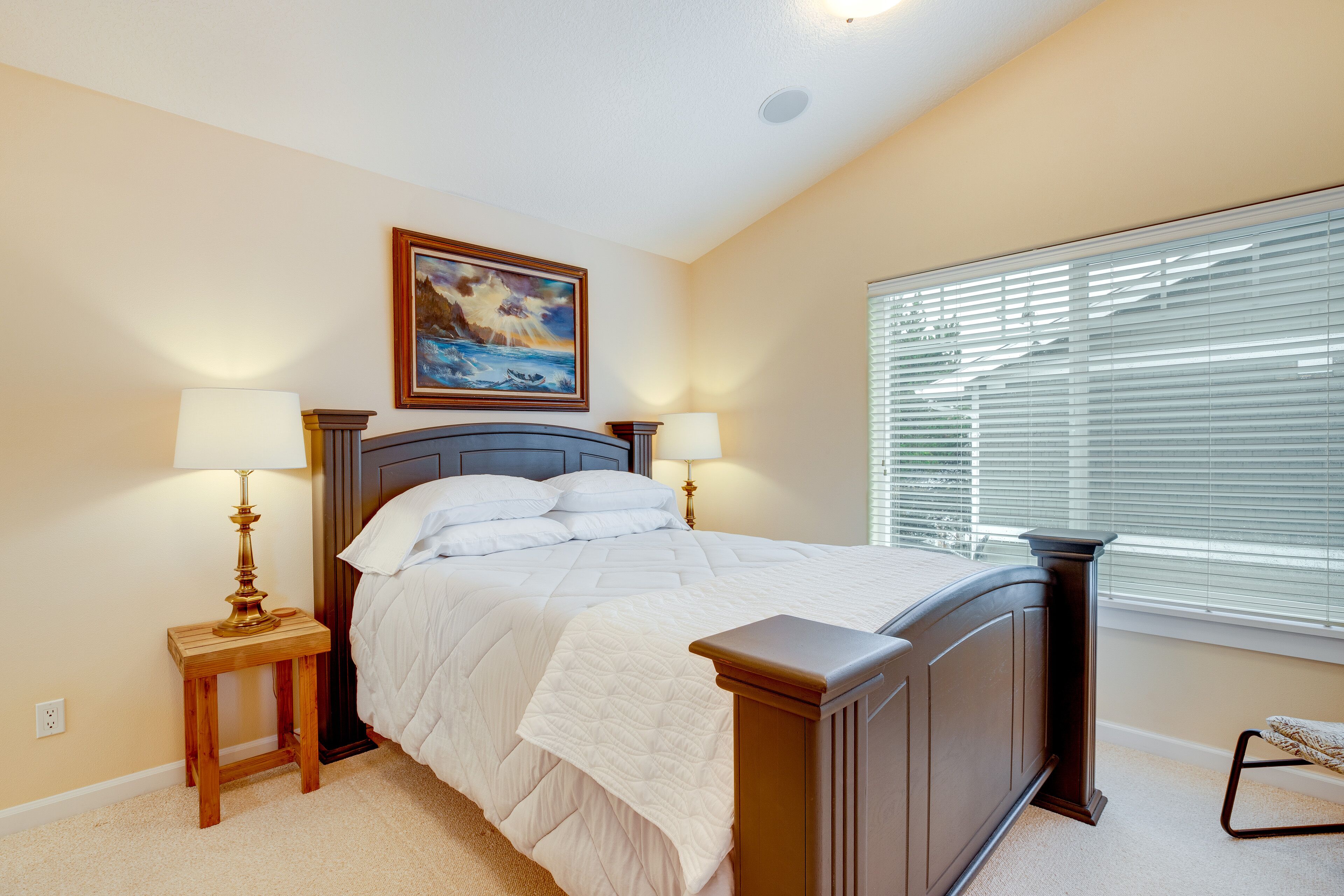 Une chambre à coucher comprenant un lit, deux tables de chevet, une commode, un tableau au-dessus du lit et une fenêtre avec des stores.
