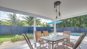 A covered patio area with a table and chairs, a hanging light fixture, and a view of a backyard with a pool and palm trees. - Chic Florida Retreat w/ Pool, Patio & Gas Grill! (Wilton Manors)