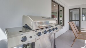 A stainless steel Char-Broil gas grill with four burners, a side burner, and a water pan, positioned next to a glass table and a chair in a modern, well-lit room with large windows. - Chic Florida Retreat w/ Pool, Patio & Gas Grill! (Wilton Manors)
