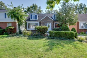 A two-story house with a brick facade, a white front door, and a small front yard with a hedge and a tree.