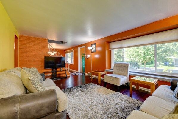 A modern living room with a brick accent wall, a flat-screen TV, a sofa, a chair, and a large window with a view of greenery.