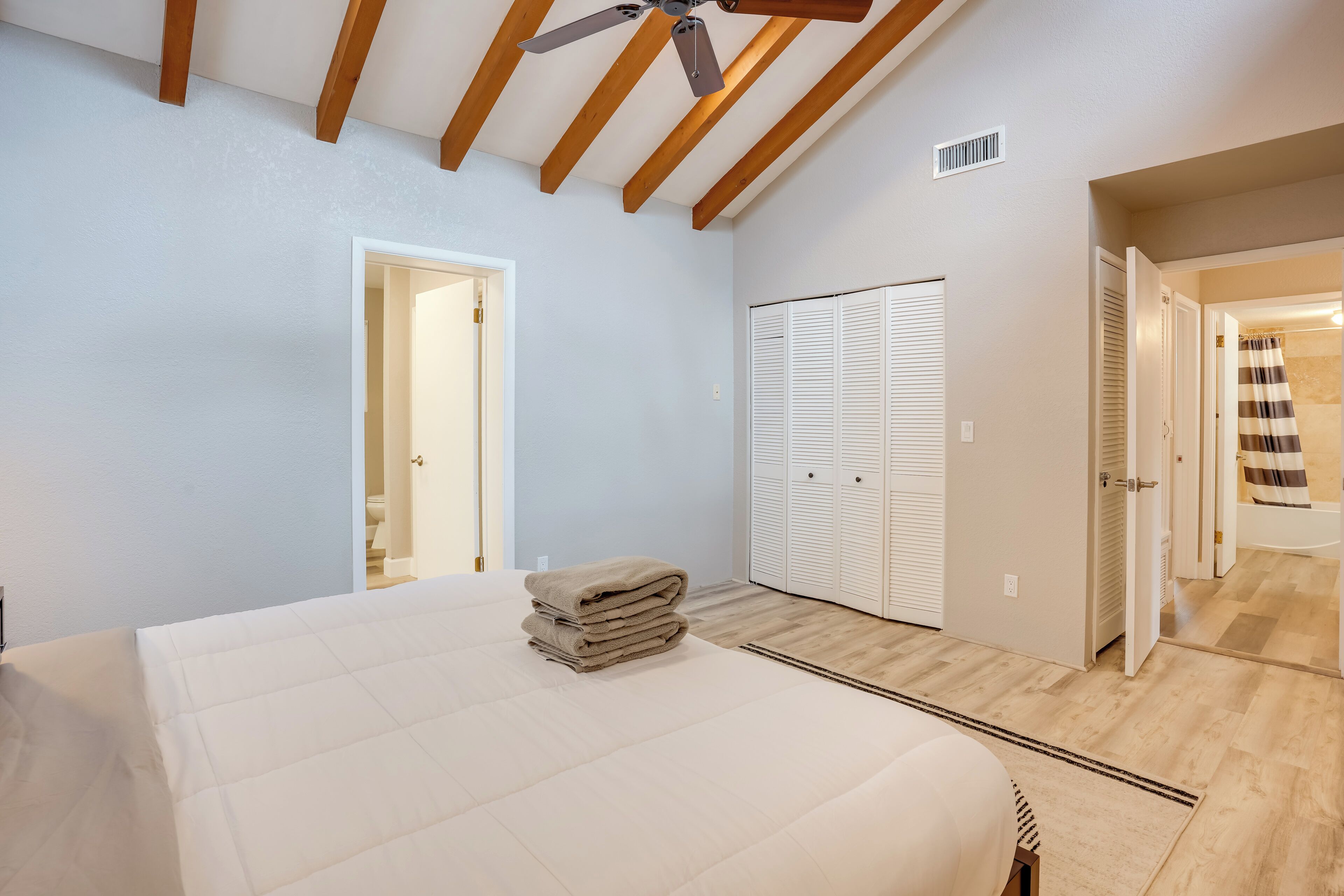 A bedroom with a bed, a ceiling fan, wooden beams, a closet, and a bathroom visible through an open door.