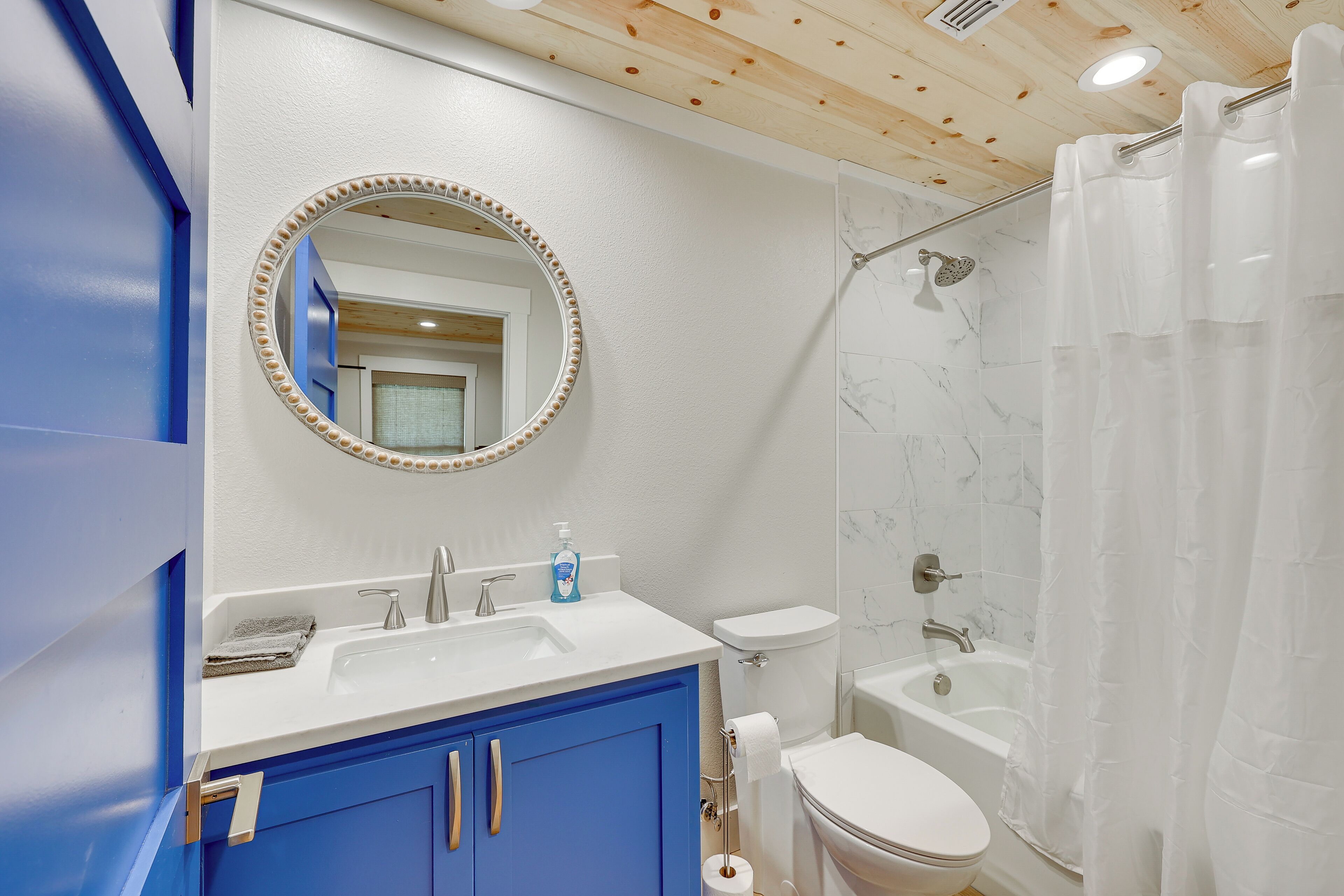 Ein Badezimmer mit einem weißen Waschbecken, blauen Schränken, einem runden Spiegel mit Perlenrahmen, einer Toilette und einer Badewanne mit Duschkopf.