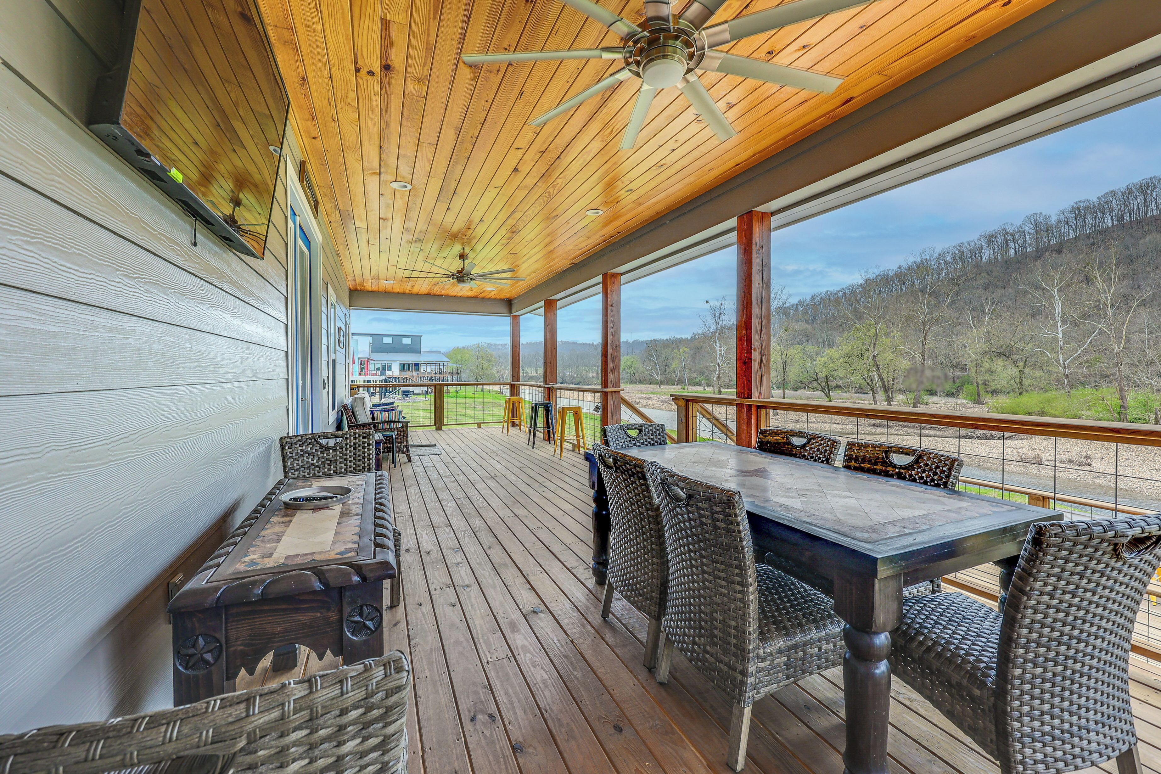 A covered deck with a table and chairs, wooden ceiling, and a view of a house and landscape.