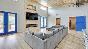 A spacious living room with a large grey sectional sofa, a fireplace, and a ceiling fan. - Riverfront 'caddo Shack' Home w/ Views & Fire Pit (Glenwood)