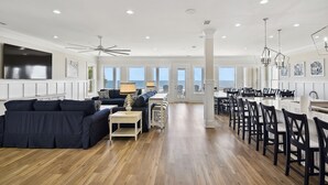 A spacious living room with a dark blue sectional sofa, a dining area with a long table and chairs, and a large TV mounted on the wall.