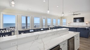 A modern kitchen with a large island, white marble countertops, and pendant lighting. There are multiple windows offering ocean views.
