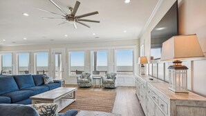 Un salón amplio con un sofá azul grande, una mesa de centro y un ventilador de techo. Hay dos sillones y una mesita con una lámpara. La habitación tiene ventanas grandes y vistas al océano.