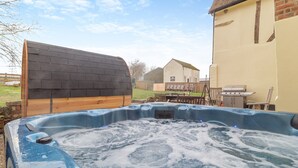 Un jacuzzi en un patio trasero con terraza de madera, una parrilla de barbacoa y una casa al fondo.