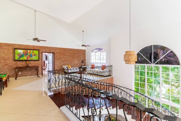 A spacious living area with a high ceiling, a fireplace, a billiards table, and a balcony with a view of greenery.