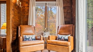 A cozy cabin interior with two leather armchairs, a wooden floor, a window with a view of autumn trees, and a lamp on a wooden side table. - Winter Wonderland of fun near Attractions (Sevierville)