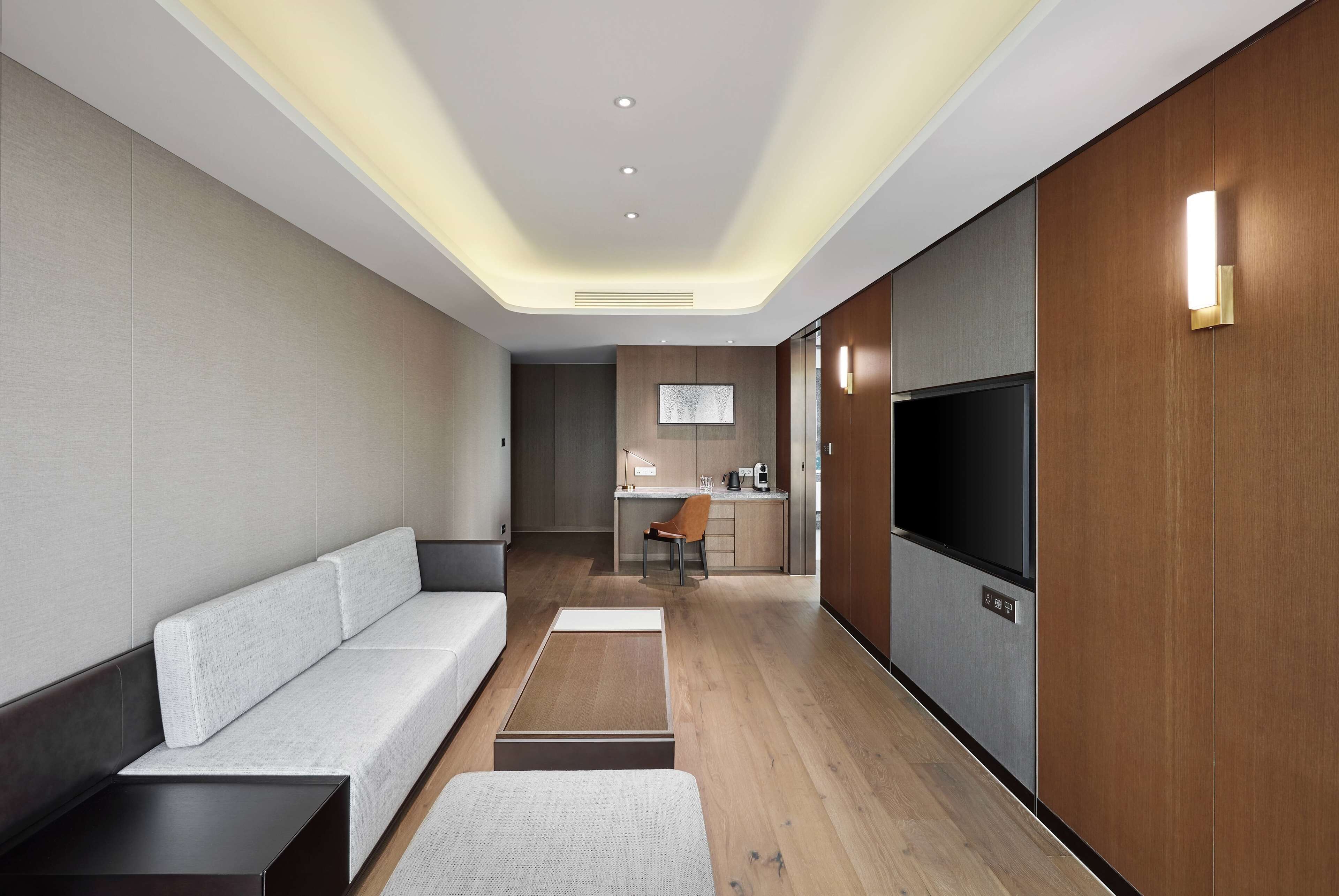 A modern hotel room with a sofa, a small table, a TV, and a kitchenette in the background.