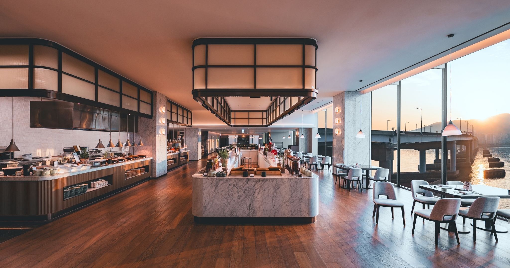 A modern restaurant with a central island, wooden flooring, and large windows overlooking a body of water.