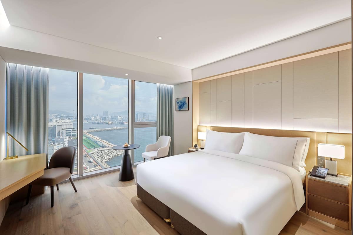 Ein Hotelzimmer mit einem großen Bett, einem Schreibtisch, einem Sessel, einem kleinen Tisch und Blick auf eine Stadtlandschaft durch große Fenster.