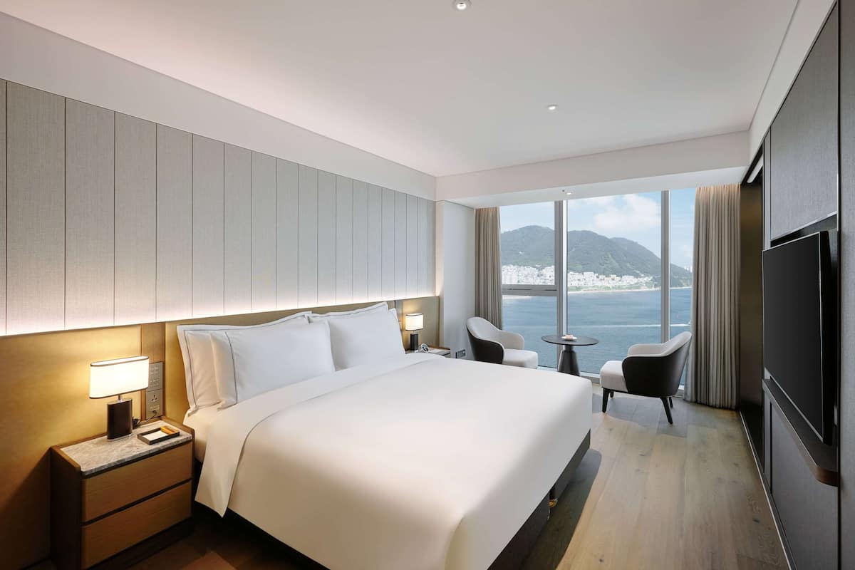 Ein modernes Hotelzimmer mit einem großen Bett, Nachttischen mit Lampen, einem Fernseher, einer Sitzecke mit Sesseln und Blick auf das Meer und die Berge durch große Fenster.