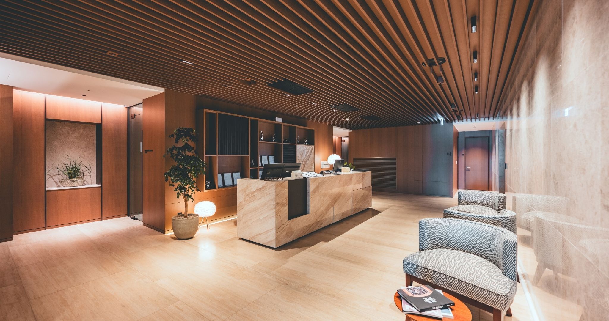 Un espace d’accueil moderne, doté d’un comptoir de réception en bois, d’un coin salon et d’une grande plante en pot.