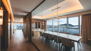 A modern hotel room with a dining area, large windows with a city view, and a glass partition.