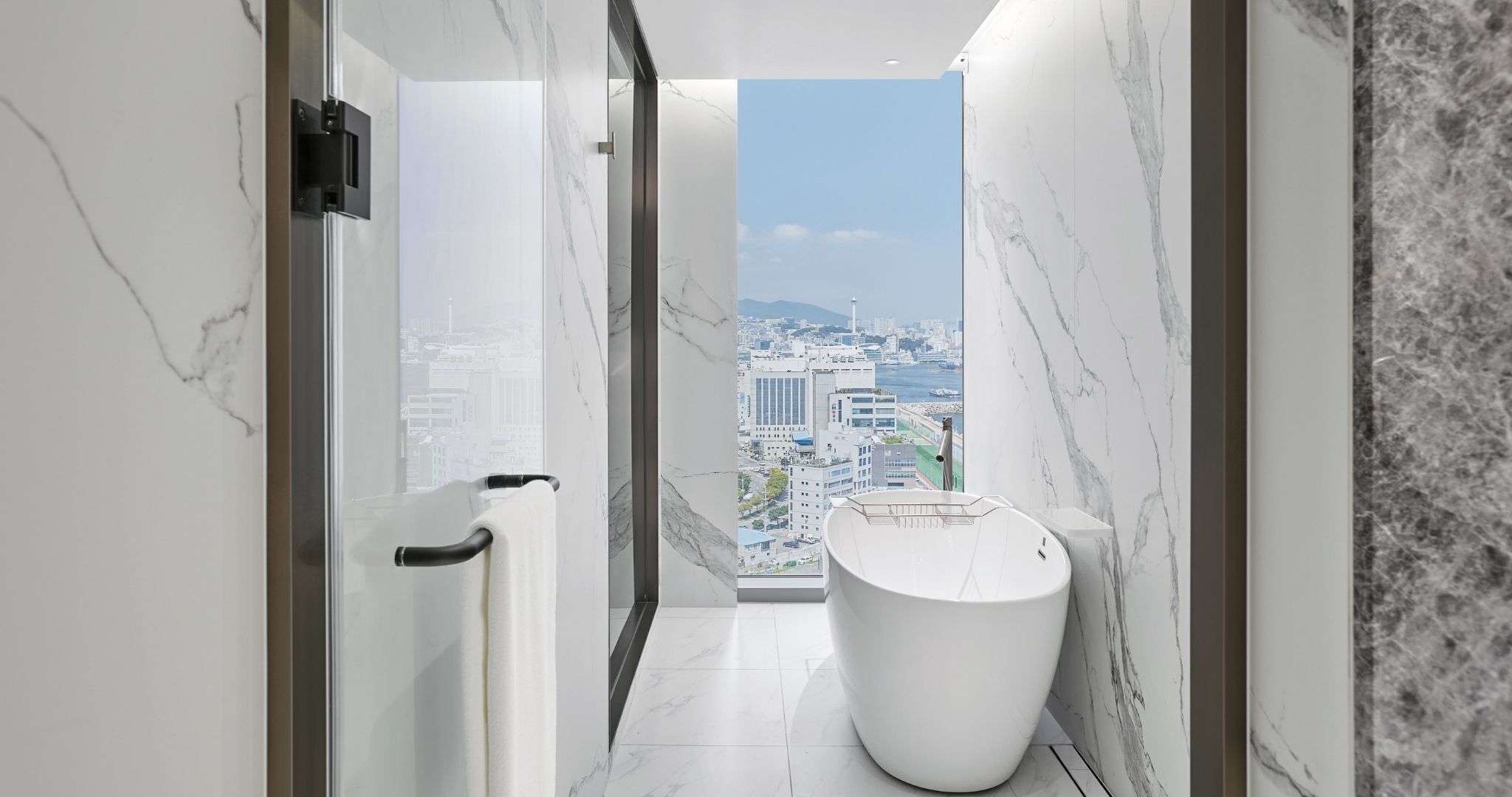 A modern bathroom with a bathtub, a view of a cityscape through a large window, and marble walls.