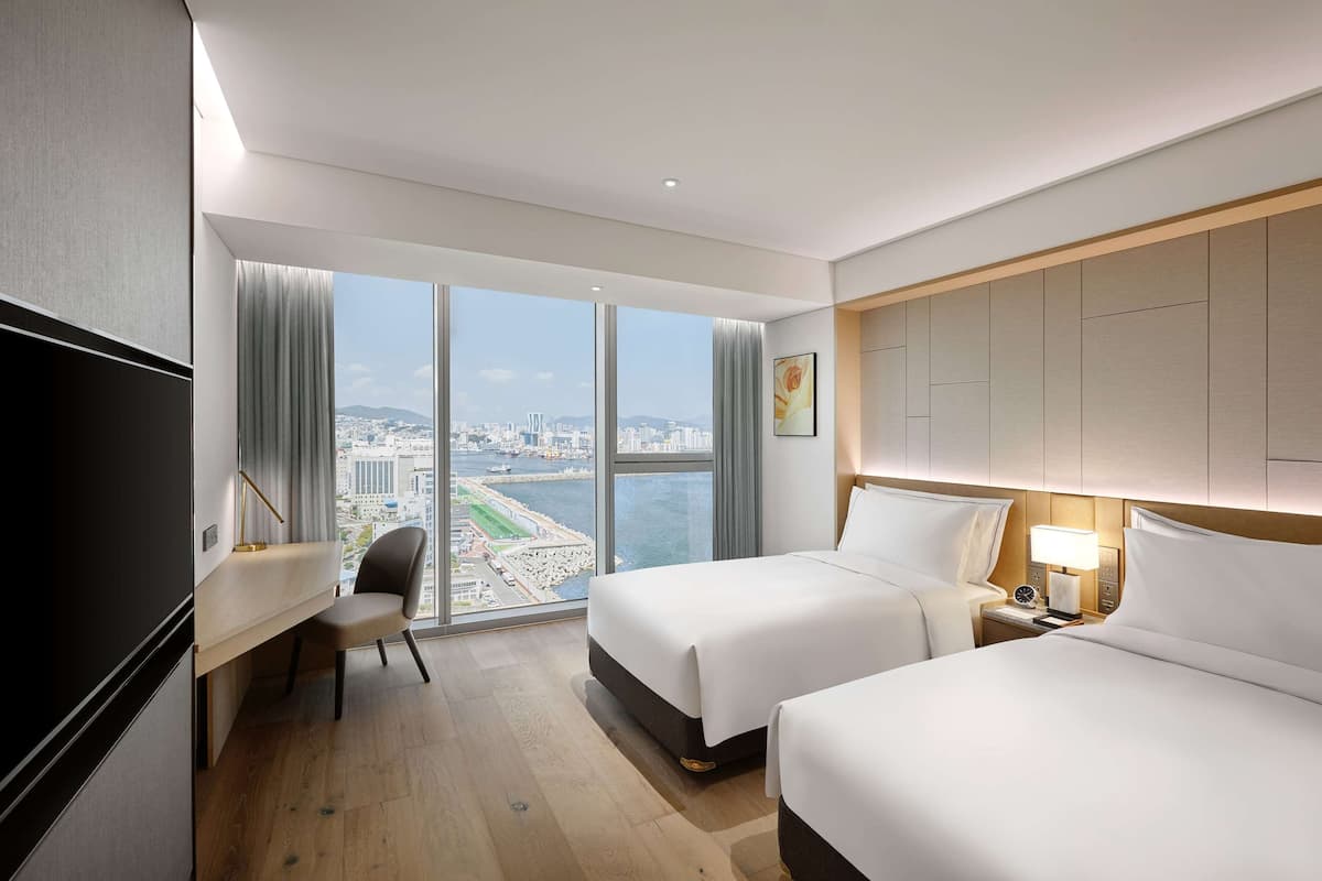 Ein Hotelzimmer mit zwei Betten, einem Schreibtisch, einem Stuhl und einem großen Fenster mit Blick auf die Stadt.