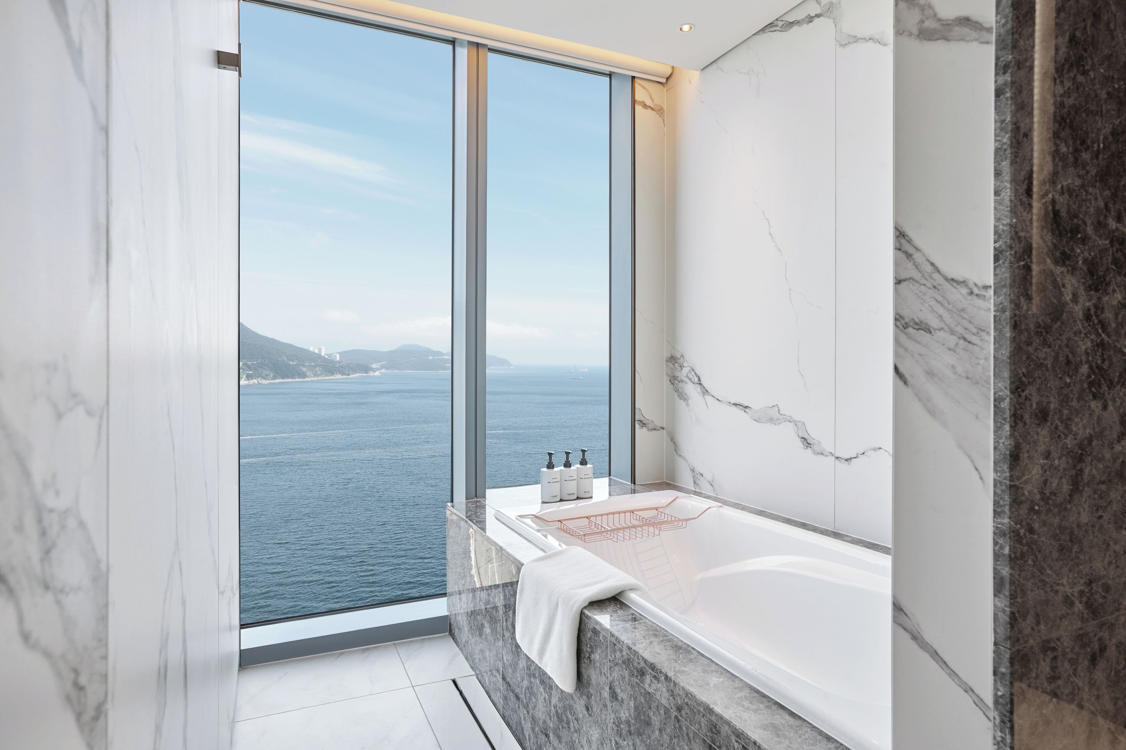 A modern bathroom with a large bathtub, marble walls, and a view of the sea.