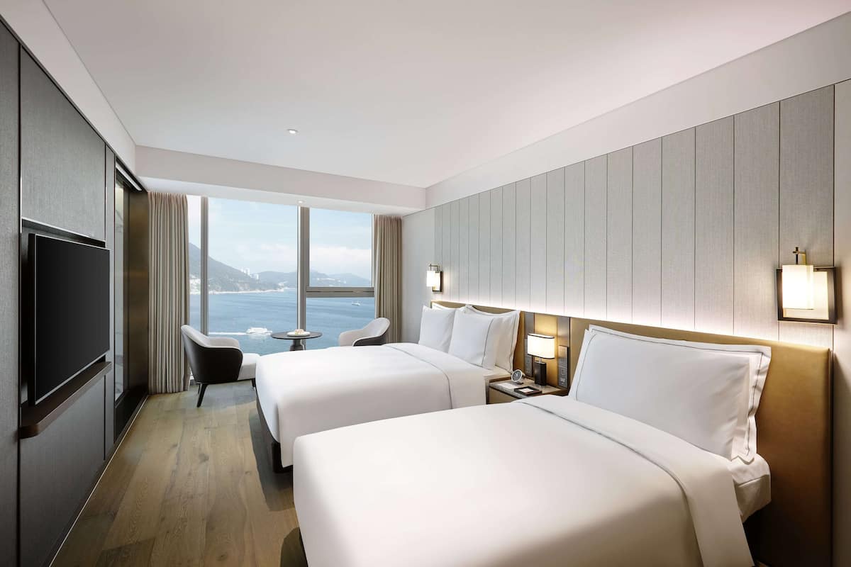 Ein Hotelzimmer mit zwei Betten, einem Fernseher, einem Schreibtisch und Meerblick.